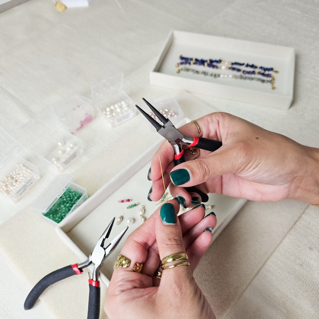 Les mains de Sahra, créatrice de Merveilles en Soi, travaillant une tige dorée au dessus d'un plateau de perles d'eau douce et de pierres naturelles